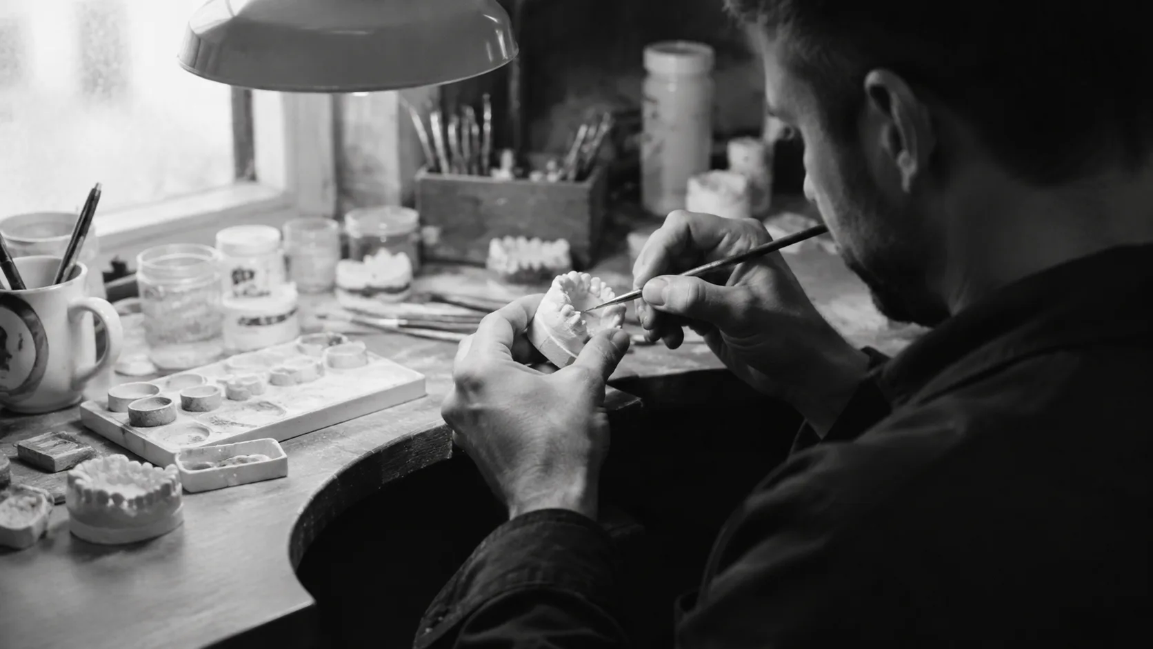 Illustrative ceramist hands working on a dental model with a fine brush.